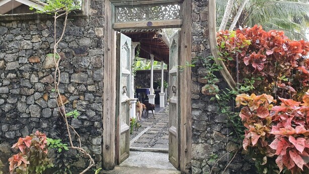 A stone archway leading to a covered area with a stone wall on one side and a garden with red foliage on the other.