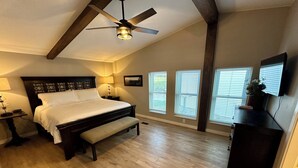 Une chambre avec un grand lit, un banc, un ventilateur de plafond et plusieurs fenêtres équipées de stores.