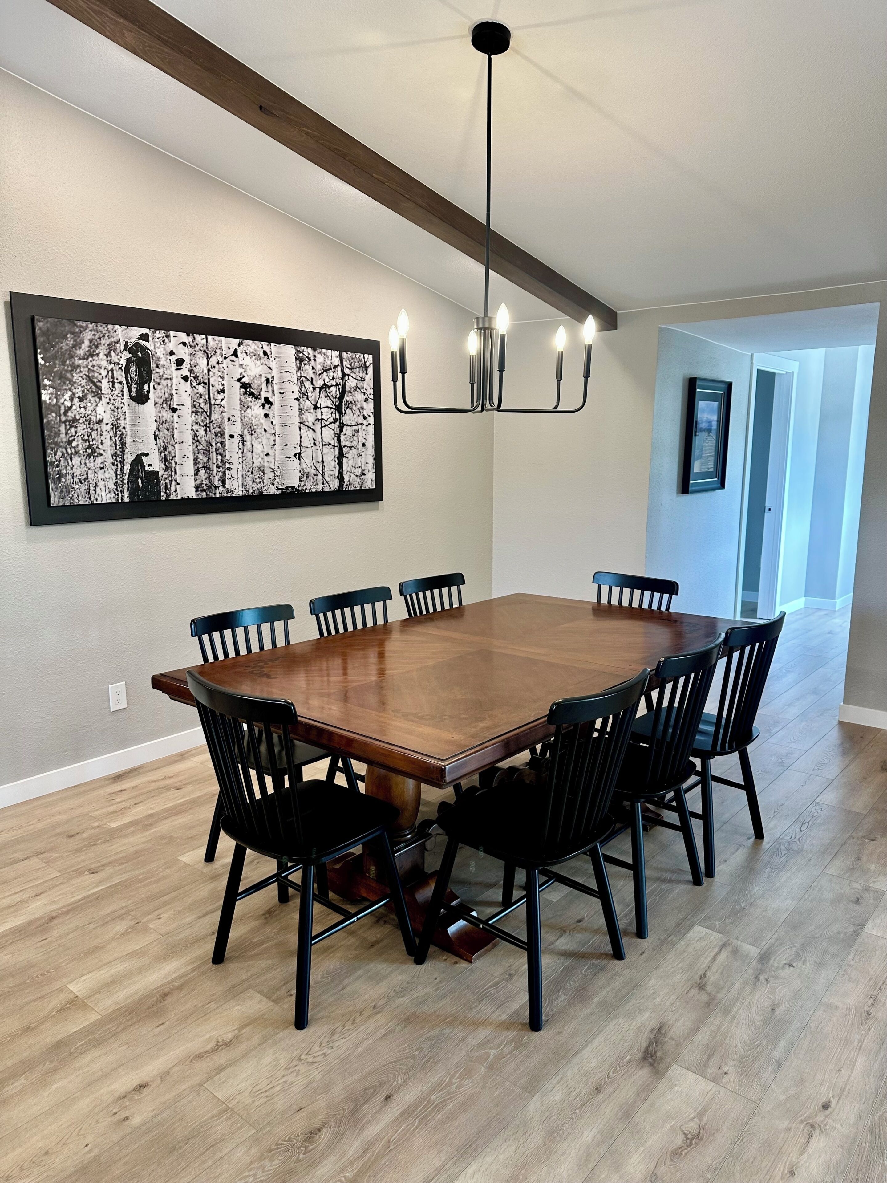 A dining area with a large wooden table surrounded by chairs, a chandelier, and a framed black and white photograph on the wall.