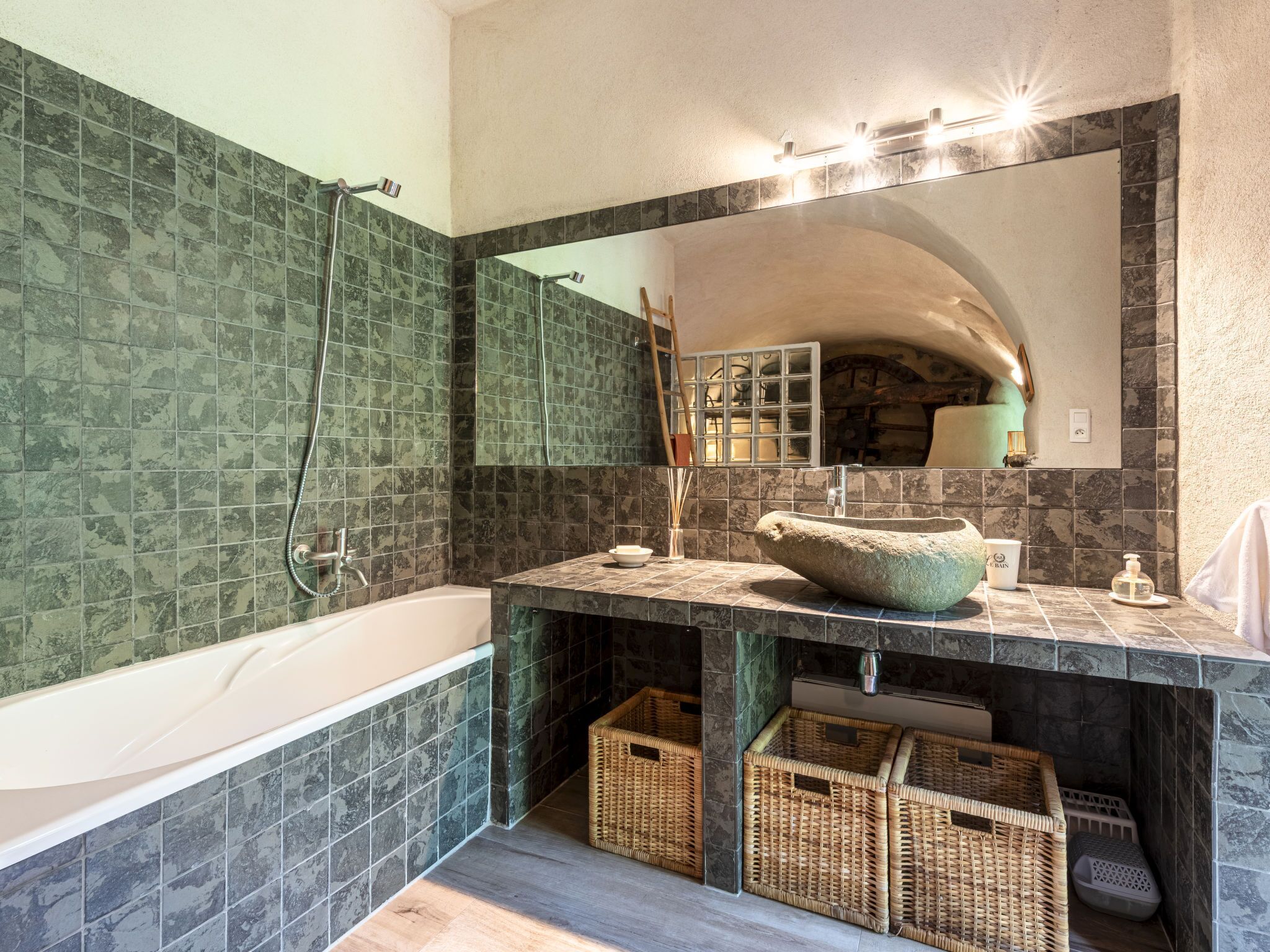 A bathroom with a bathtub, a large mirror, and a sink with a stone basin. There are wicker baskets under the sink and a towel hanging on the wall.