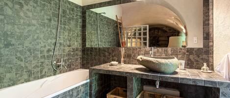 A bathroom with a bathtub, a large mirror, and a sink with a stone basin. There are wicker baskets under the sink and a towel hanging on the wall.
