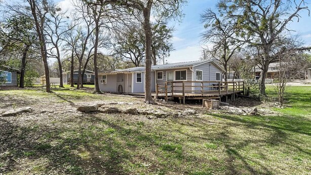 A single-story mobile home with a porch and a small deck, surrounded by trees and a grassy yard.