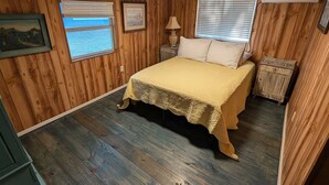 A bedroom with wooden paneling, a bed with a yellow bedspread, two windows with blinds, a nightstand with a lamp, and a small wooden chest.