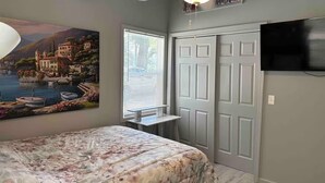 A bedroom with a bed, a ceiling fan, a painting of a lakeside scene, a window with blinds, and a TV mounted on the wall.