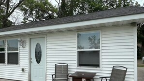 A small, single-story building with a white exterior and a dark brown door. There are two windows with blinds, a small porch area with a metal table and two chairs, and a doormat in front of the door.