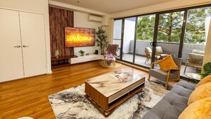 Un salón moderno con suelo de madera, una mesa de centro de mármol y un sofá. Hay un ventanal con vistas a árboles y un balcón con mobiliario de exterior.