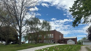 Tiilestä rakennettu rakennus, jonka edessä on suuri puu, soralla päällystetty polku ja nurmialue, jossa kasvaa kukkia.