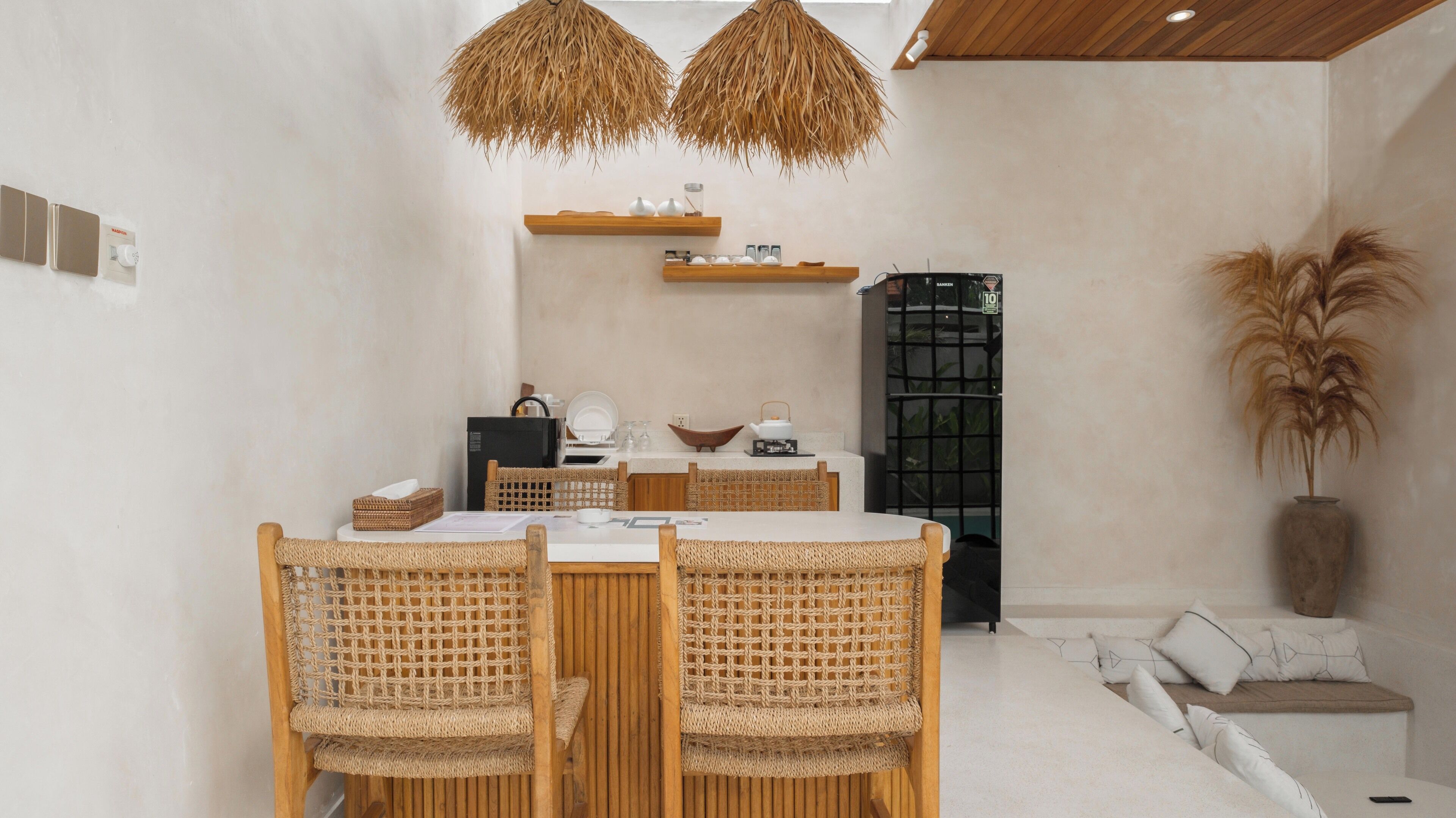 Comedor con una mesa redonda y dos sillas de mimbre, un refrigerador negro y una estantería con objetos decorativos.