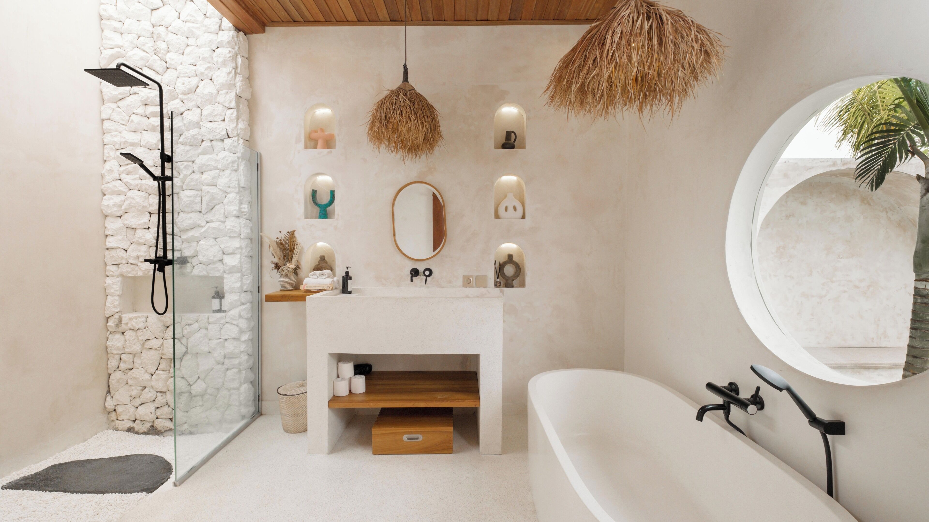 A modern bathroom with a stone wall, a freestanding shower, a vanity with a mirror, and a round mirror on the wall.