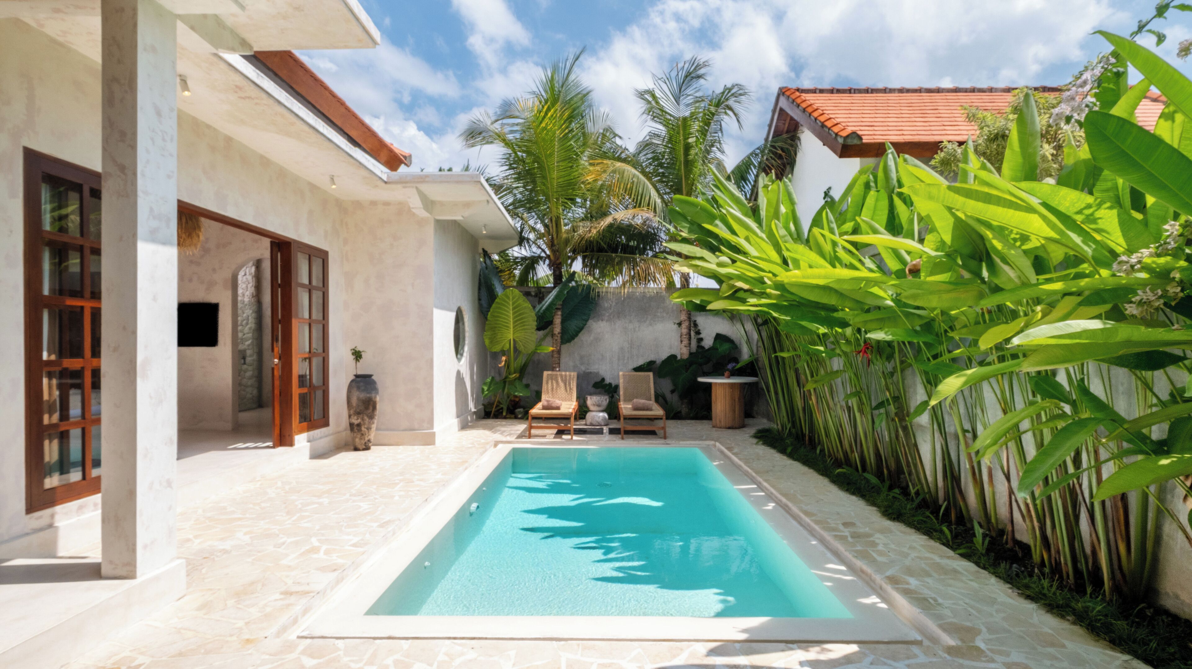 A swimming pool with two lounge chairs and a small table, surrounded by lush tropical plants and a stone-tiled patio.