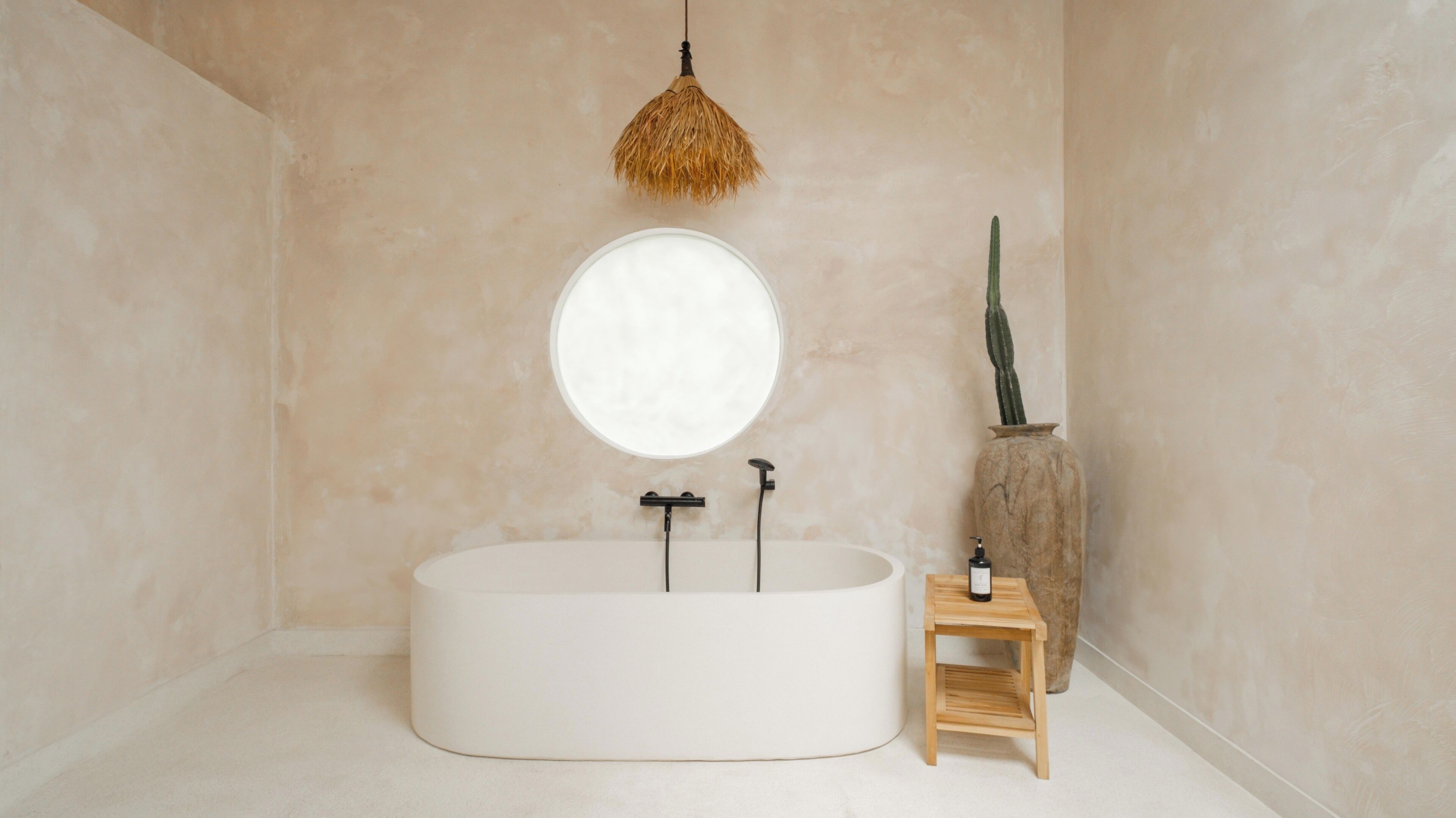 Un bagno dal design minimalista con una vasca freestanding, uno specchio rotondo, uno sgabello in legno e un cactus in vaso.