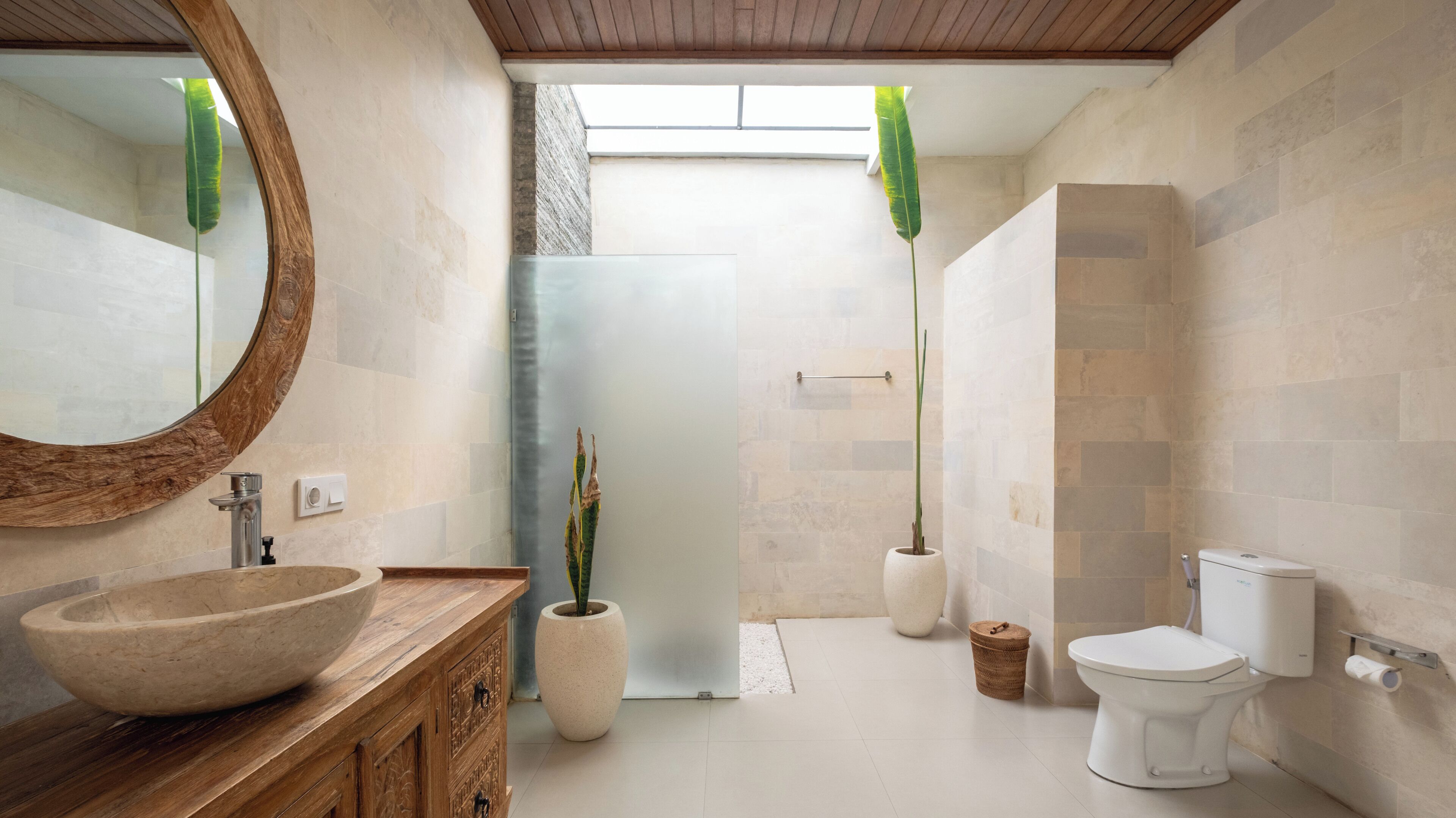 A modern bathroom with a large wooden vanity, a round mirror, a stone sink, and a toilet. There are two potted plants and a shower area visible.