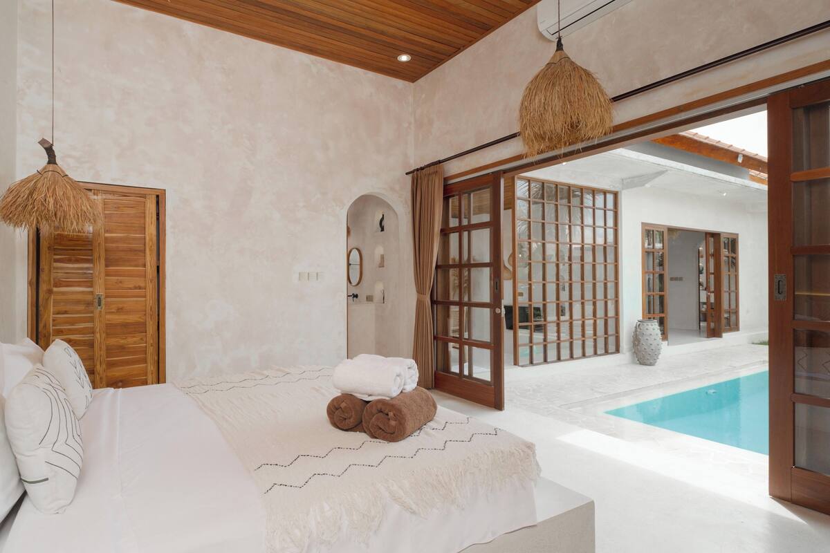A bedroom with a bed, a wooden door, a mirror, a window, and a view of a swimming pool.
