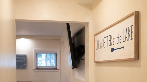 A hallway with a wooden floor, a couch, a TV mounted on the wall, and a sign that reads "Life is better at the lake". - The Nautilus at JS Hubbs Guest House (Hammondsport)