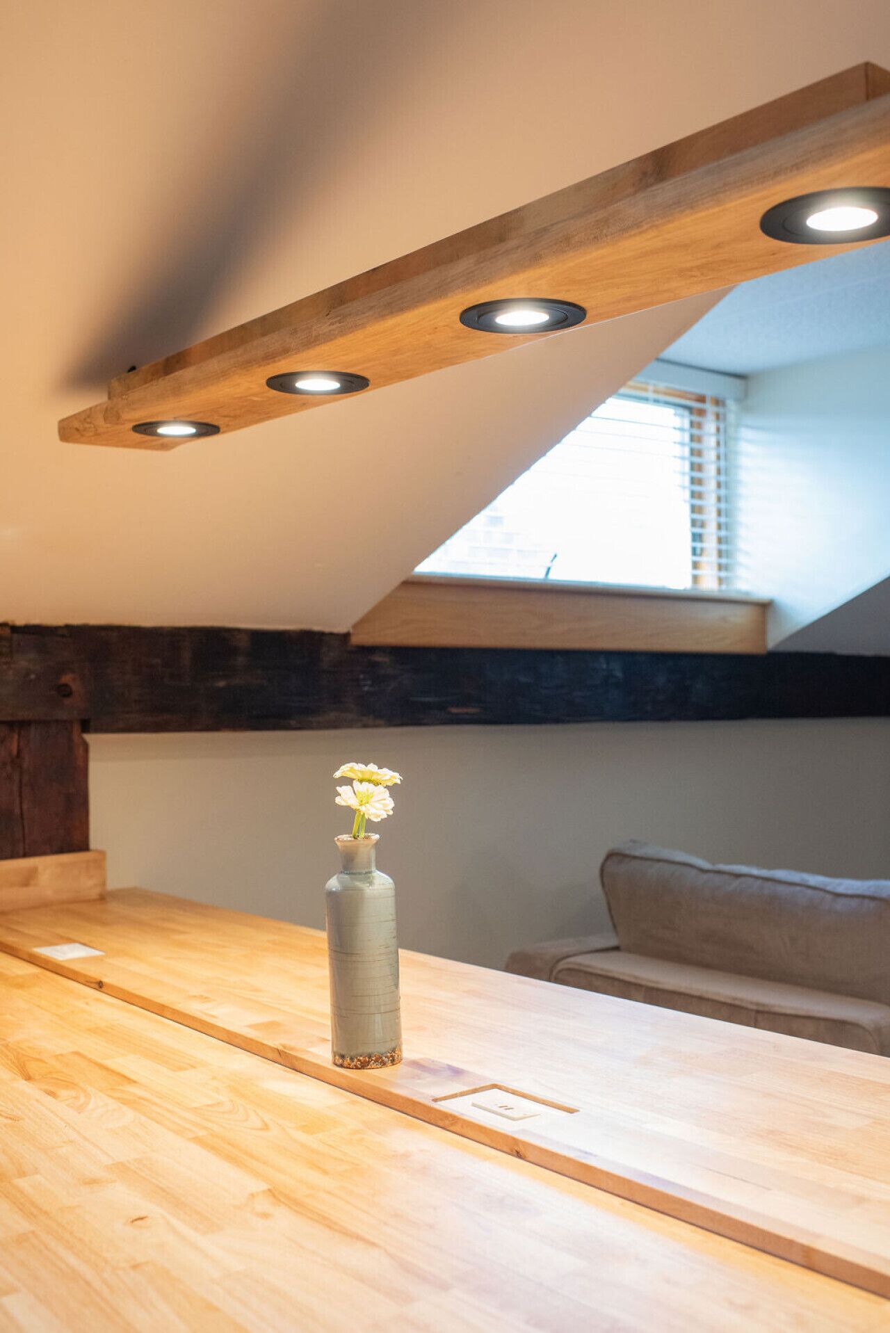 A wooden dining table with a vase of flowers, a wooden ceiling with recessed lighting, and a view of a window with blinds.