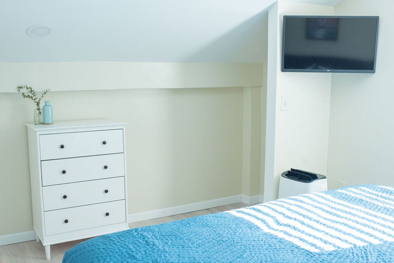 A bedroom with a white dresser, a blue and white striped bedspread, a television mounted on the wall, and a trash bin.