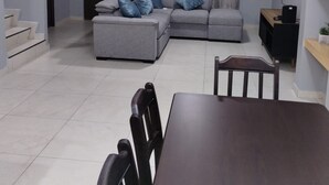 Un salon moderne avec un canapé d’angle gris, une table à manger avec des chaises, un téléviseur fixé au mur et un escalier menant à l’étage supérieur.