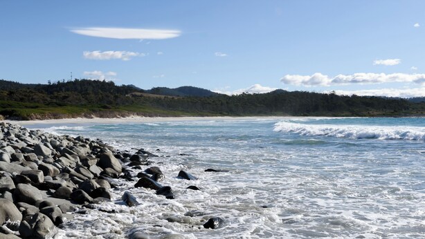 A rocky beach with waves, a clear sky, and distant hills.