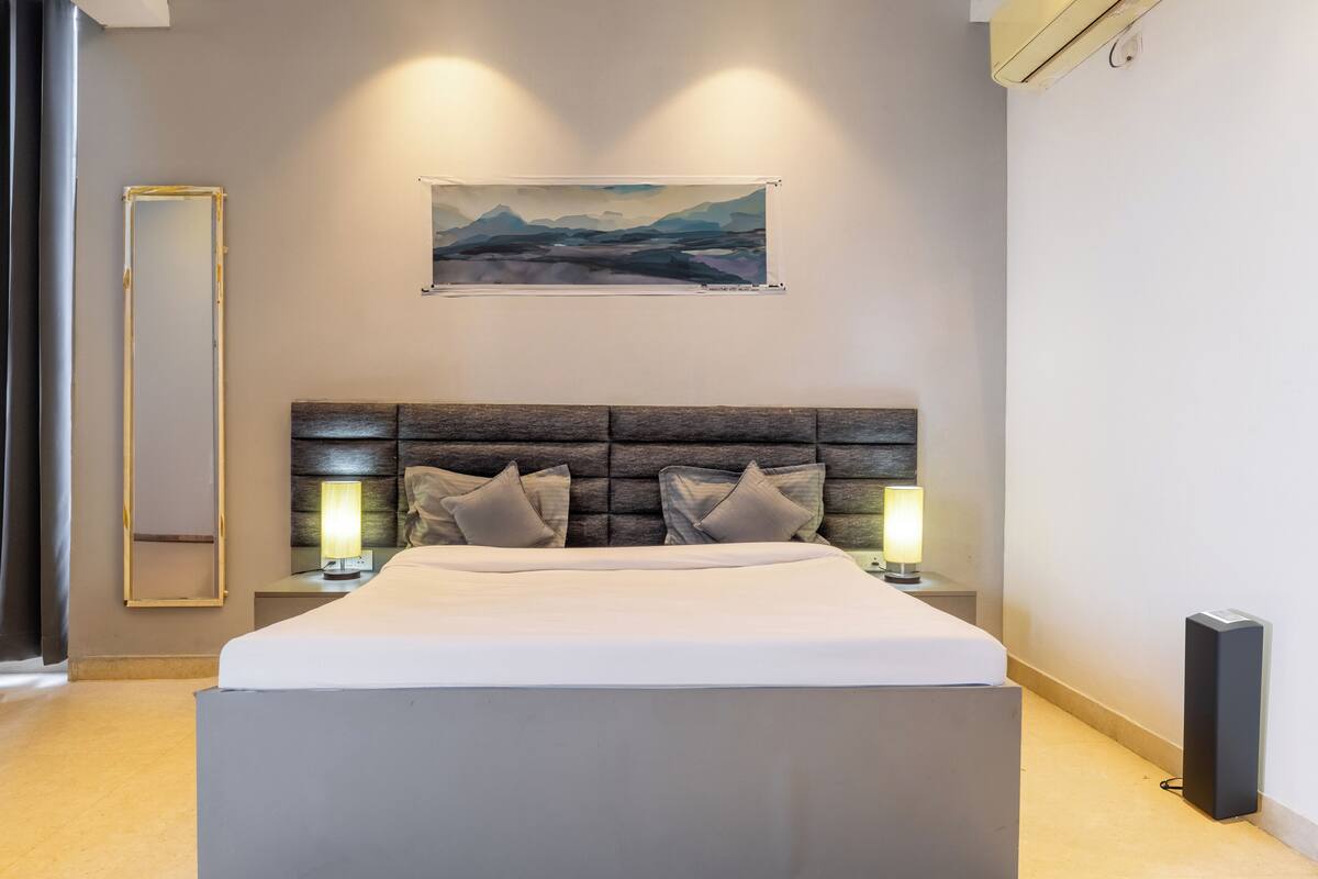 Een moderne slaapkamer met een groot bed, een hoofdbord met ingebouwde verlichting, een nachtkastje met een lamp en een lijst met een landschapsafbeelding aan de muur.