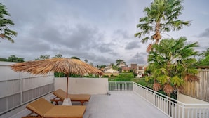 A rooftop terrace with two lounge chairs, a thatched umbrella, and a view of the neighborhood.