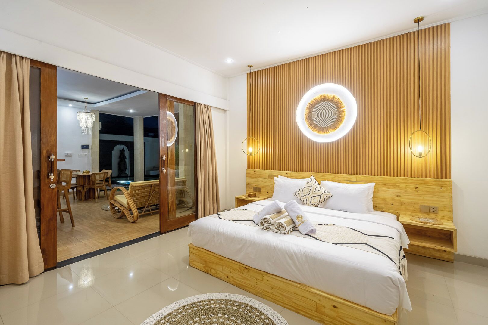 Una habitación de hotel moderna con cabecera de madera, una cama grande con ropa blanca, una lámpara de pared redonda y un área de comedor visible a través de una puerta corredera.