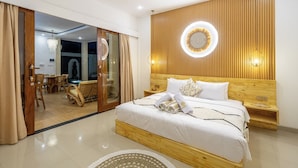 A modern hotel room with a wooden headboard, a large bed with white linens, a round wall light, and a dining area visible through a sliding door.