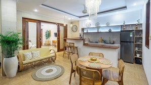 A modern living room with a sofa, coffee table, and chairs, a dining area with a table and chairs, a kitchen with a refrigerator and shelves, and a large plant.