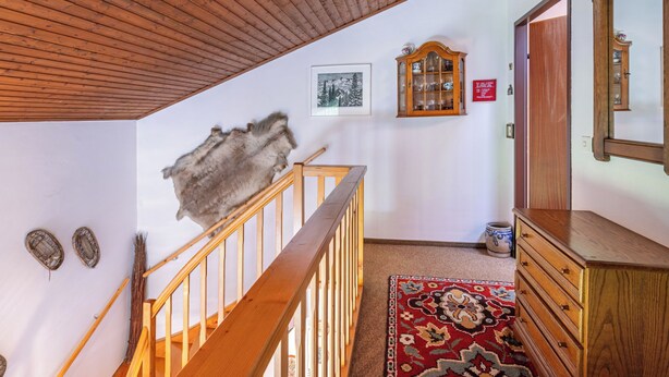 Eine Holztreppe führt zu einem Schlafbereich mit geneigter Decke, an der Wand hängt ein Fellstück, ein gerahmtes Bild, ein Holzschrank und ein Teppich mit rotem Muster sind zu sehen.