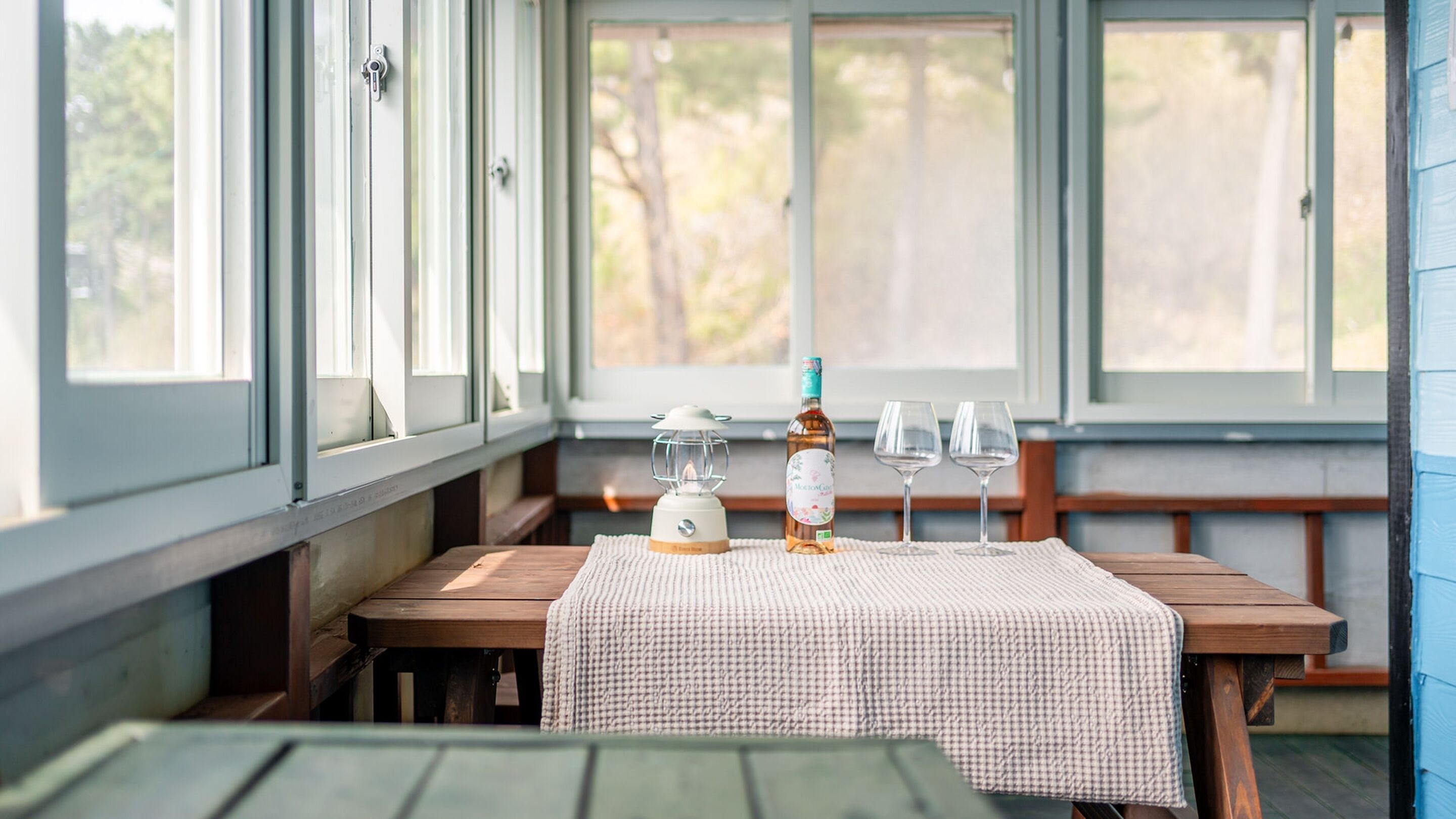 A wooden bench with a checkered tablecloth, a bottle of liquor, and two wine glasses on a table in a room with large windows.