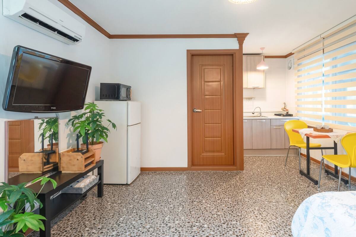 Habitació compacta amb llit, escriptori amb televisió, nevera, microones, porta, finestra amb persianes i zona de cuina amb rentapelats i cadira.