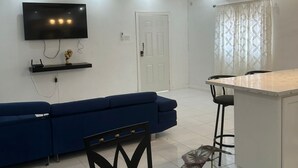 A modern living room with a flat-screen TV, a blue sofa, a dining area with a small island, and a ceiling fan.