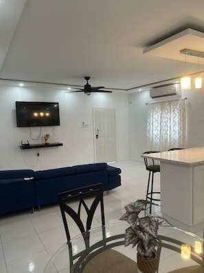 A modern living room with a flat-screen TV, a blue sofa, a dining area with a small island, and a ceiling fan.