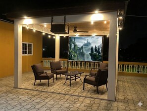 Et udendørs opholdsområde med stole og et bord, et stort skærmbillede af et fantasylandskab og en balkon med gelænder.