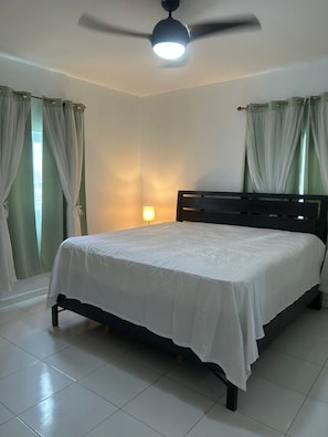 A bedroom with a bed, curtains, a ceiling fan, a wall-mounted lamp, and a tiled floor.