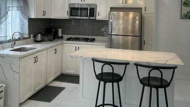 A modern kitchen with white cabinets, a stainless steel refrigerator, a microwave, and a gas stove. There are two black bar stools at the island.