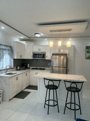 A modern kitchen with white cabinets, a stainless steel refrigerator, a microwave, and a gas stove. There are two black bar stools at the island.