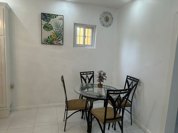 A dining area with a glass table, four chairs, a small window, and a clock on the wall.