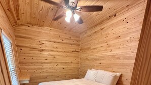 A wooden-paneled room with a bed, a ceiling fan, and a window with blinds. - Charming Pondview Cabin (Andalusia)