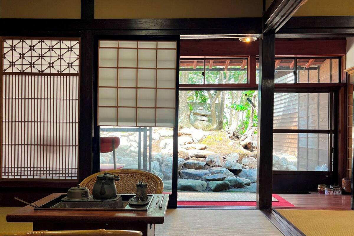 A traditional Japanese room with tatami flooring, a shoji screen, a tatami mat table, a low table with a tea set, and a view of a garden through a sliding door.