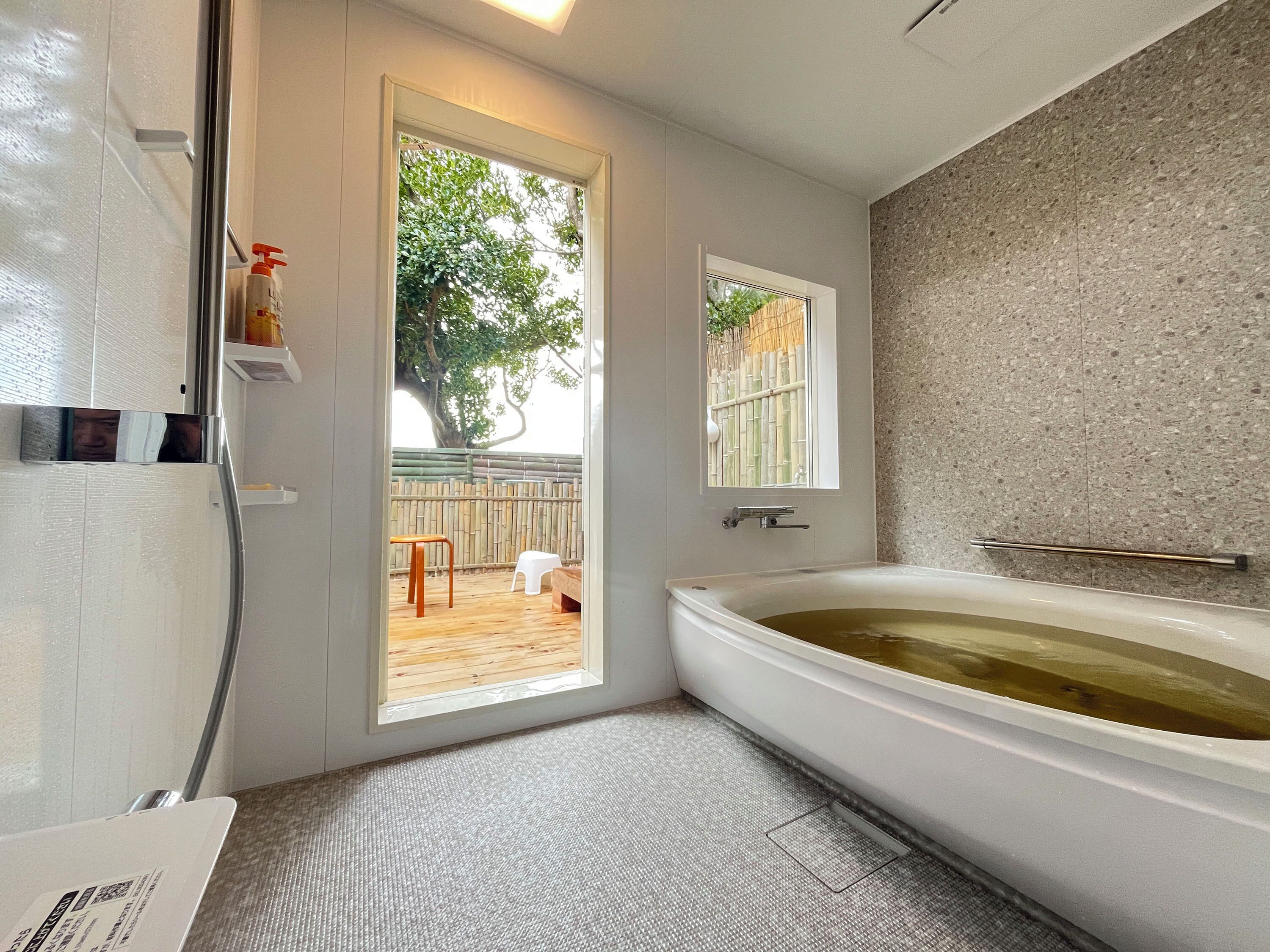 Un baño con bañera, ducha, inodoro y ventana que ofrece vistas a una terraza de madera y un árbol.