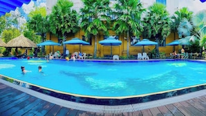 A swimming pool with lounge chairs and umbrellas, surrounded by palm trees and a yellow building.