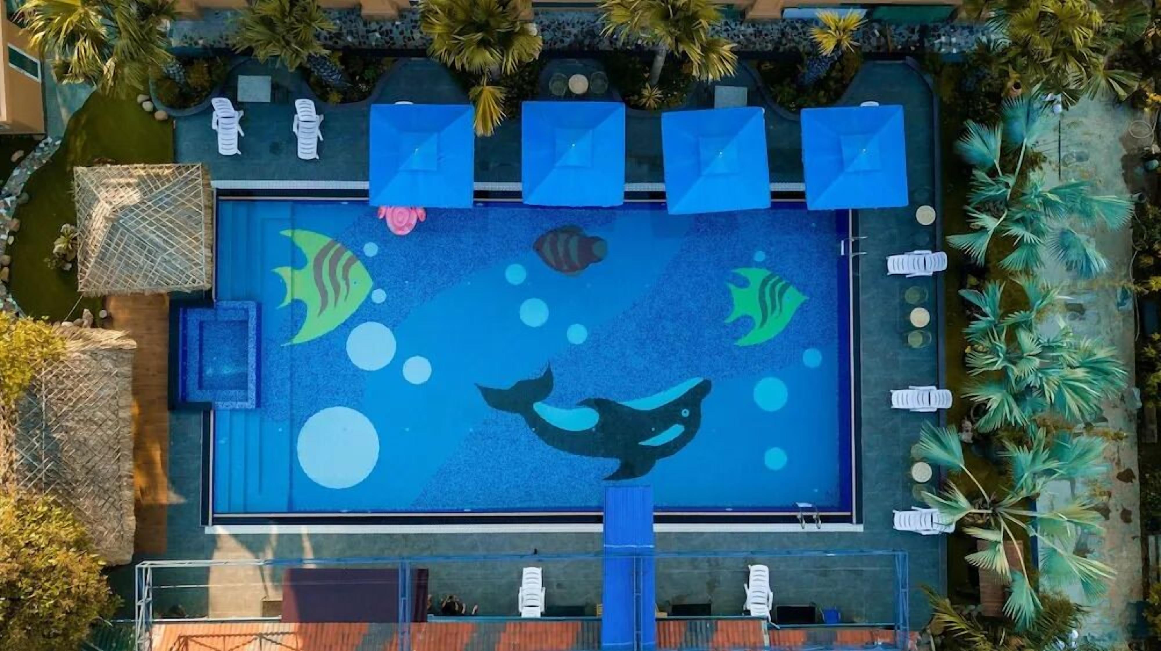 An aerial view of a swimming pool with a mural of fish and bubbles, surrounded by lounge chairs and palm trees.