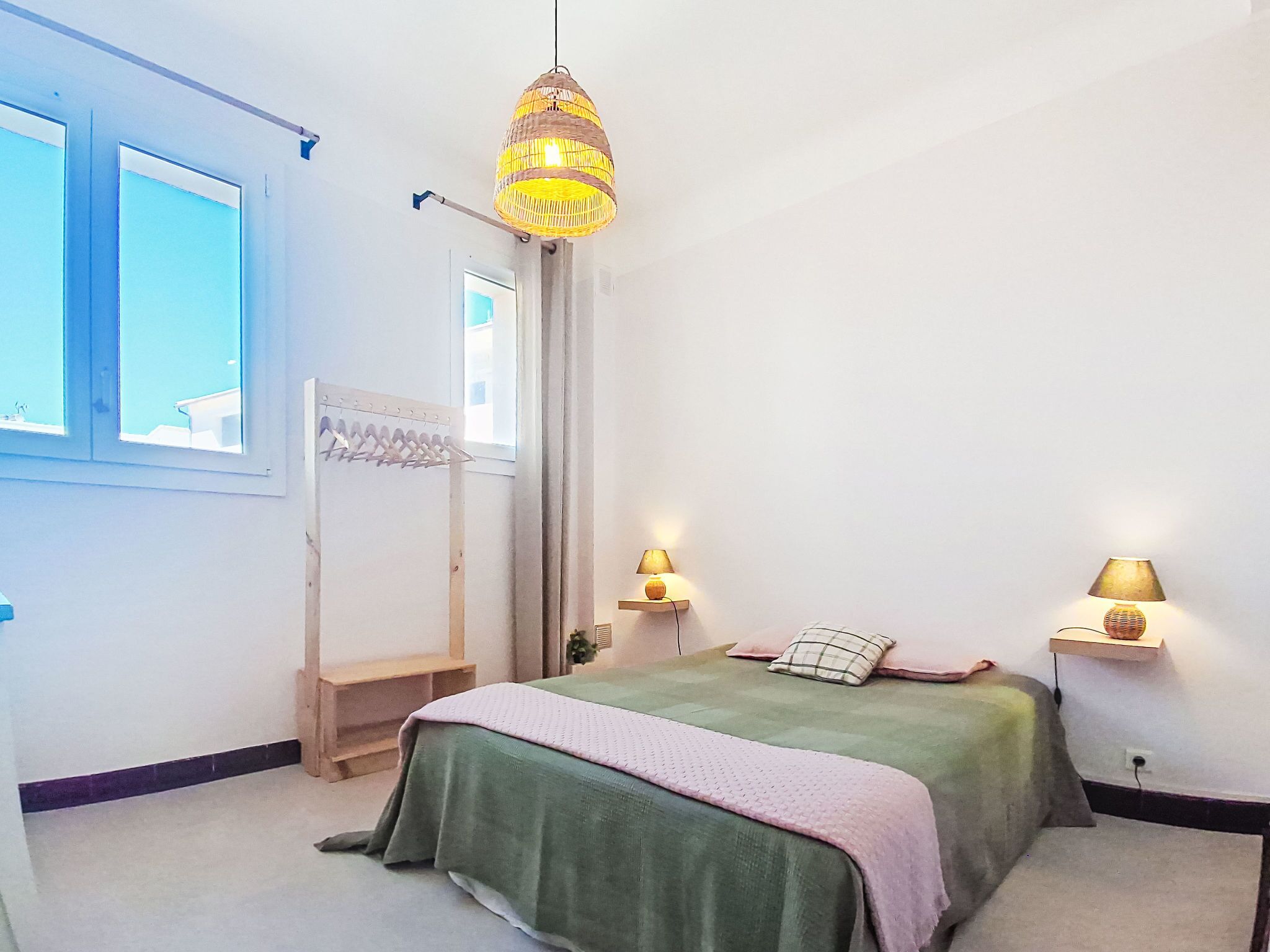 A bedroom with a bed, two bedside lamps, a hanging light fixture, a window, and a clothes rack.
