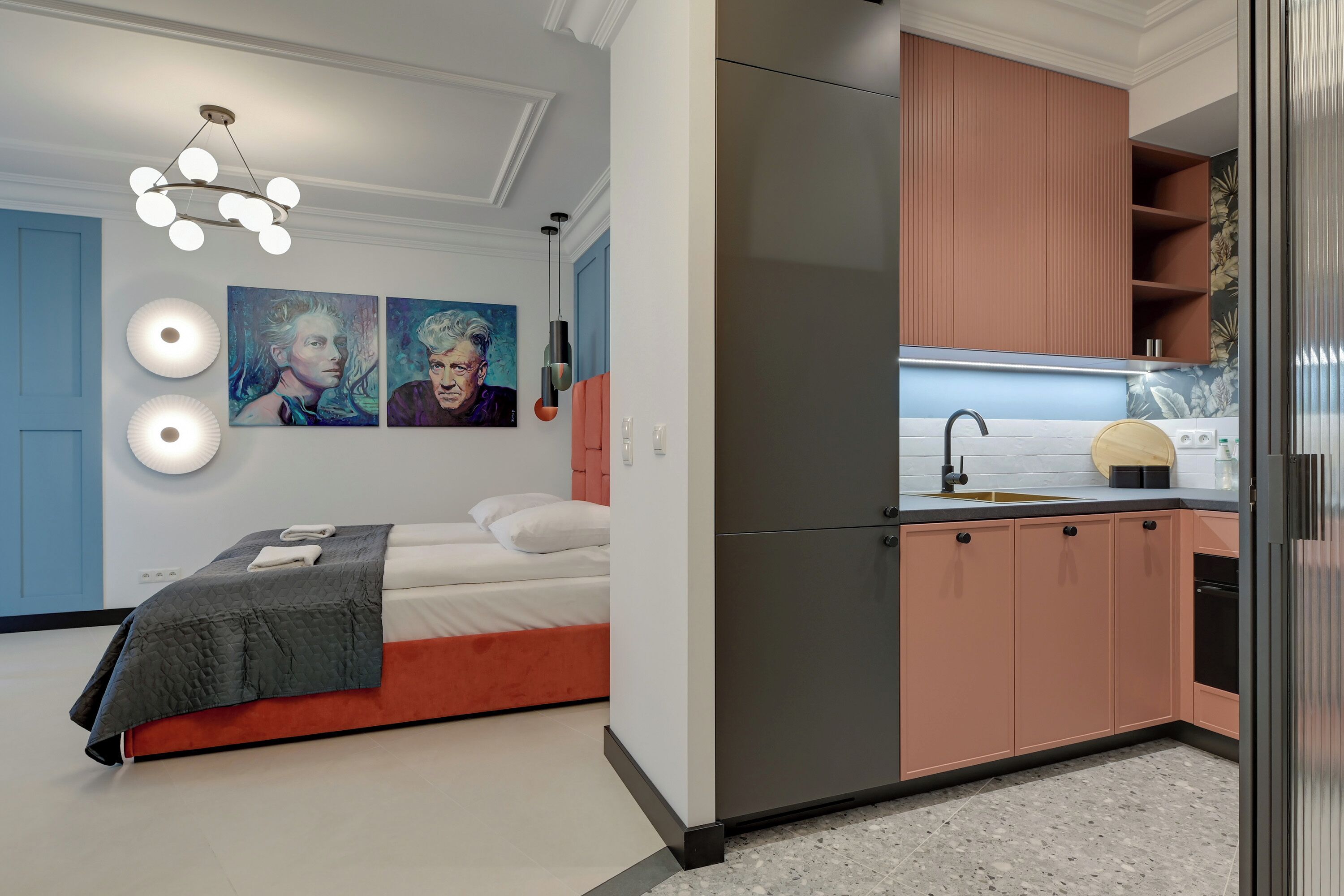 Quarto moderno com cama, mesa de cabeceira e arte na parede. Ao lado, uma cozinha compacta com geladeira, pia e armários.