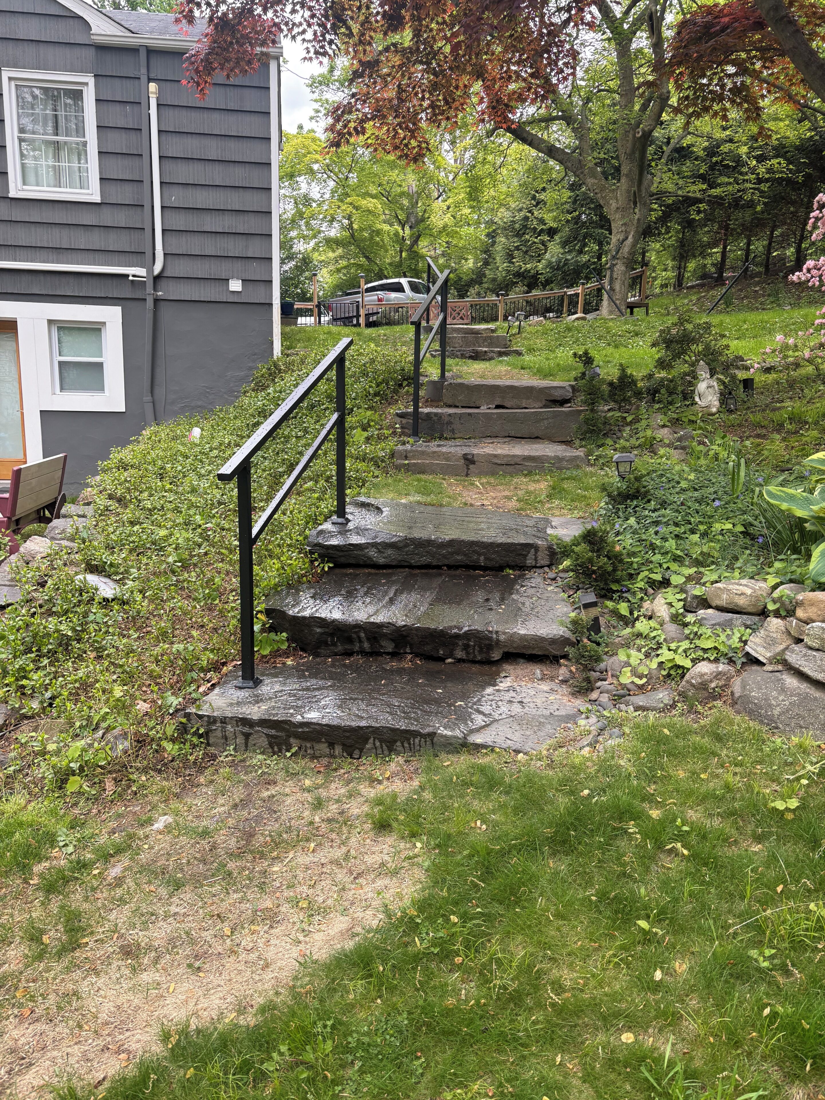 A set of stone steps leading up to a house with a black metal handrail, surrounded by greenery and a tree with autumn leaves.