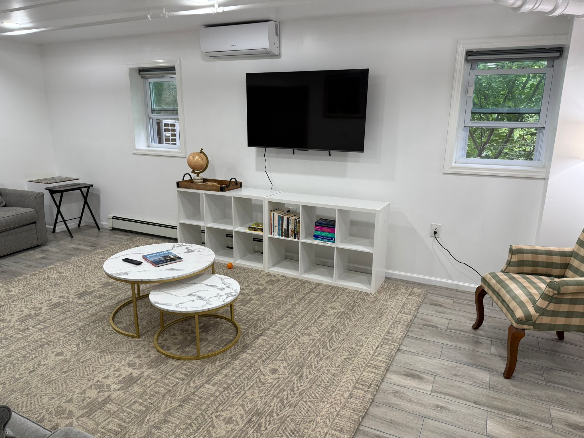 Een woonkamer met een flatscreen televisie aan de muur, een witte dressoir met boeken, een grijze bank, een ronde salontafel en een kamerkleed met dessin.