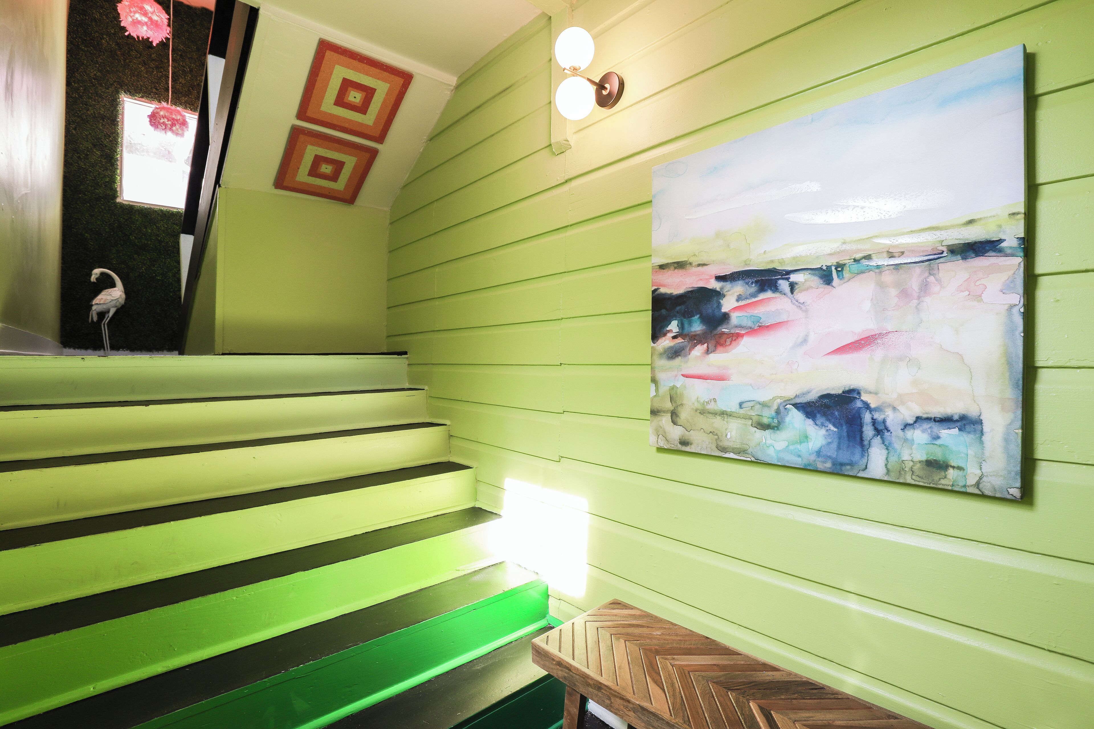 Uma escada com degraus verdes, um quadro abstrato fixado na parede e uma pequena mesa de madeira.