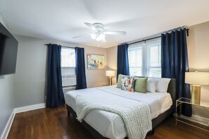 A bedroom with a large bed, a ceiling fan, two windows with curtains, a painting on the wall, and a bedside lamp. - 2BR The Green Plex! D! Downtown, Medical Center ★★★★★ (Houston)