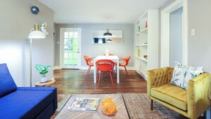 Een woonkamer met een blauwe bank, een gele fauteuil, een eethoek met een witte tafel en oranje stoelen, en een boekenkast.