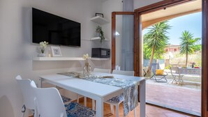 A dining area with a white table and chairs, a flat-screen TV mounted on the wall, and a view of the outdoor patio with palm trees.